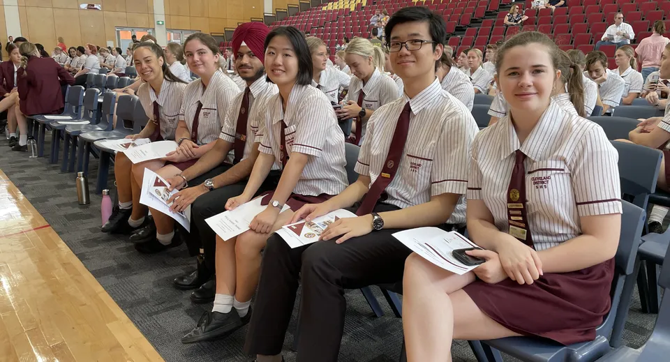 Un gruppo di studentesse della Cleveland District State High School siede in fila con lo sguardo rivolto verso un palco e posti per il pubblico sullo sfondo.