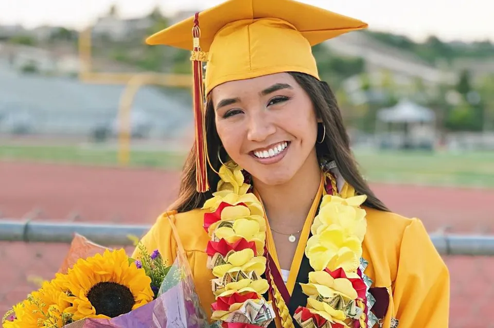 Una diplomata della Esperanza High School indossa una toga gialla e tiene in mano un bouquet di fiori davanti a un campo sportivo.