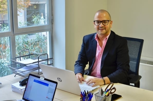 Hombre con gafas y americana sentado en una oficina trabajando con un ordenador portátil