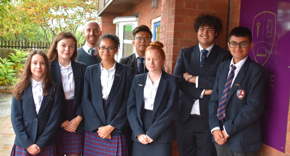 Un gruppo di studenti e studentesse della Chase Grammar School si trova insieme davanti a un edificio in mattoni con piante sullo sfondo.