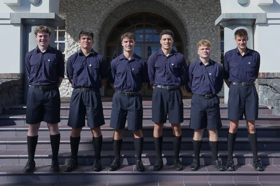 Un gruppo di sei studenti in divisa blu si trova davanti a un edificio in pietra con finestre ad arco nel campus della Auckland Grammar School.