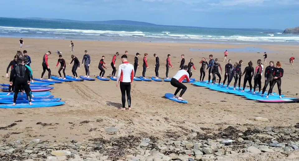 Un gruppo di studenti del Hazelwood College in muta sta sulla spiaggia con le tavole da surf davanti all'oceano.