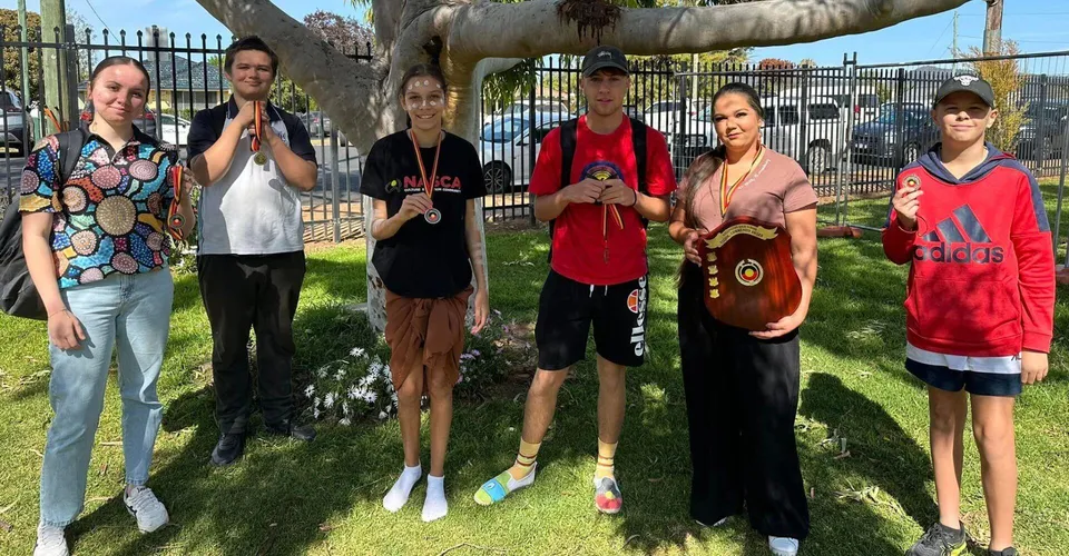 Un gruppo di studenti e adulti è in piedi su un prato all'interno del terreno della Wellington High School, davanti a un grande albero.