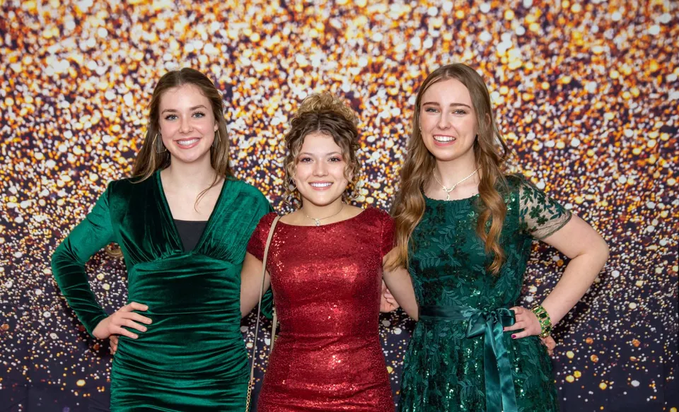 Tre studentesse della American Heritage Academy in abiti da sera sorridono davanti a uno sfondo luccicante.