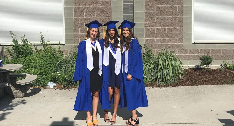 Tre studentesse della Rutland Senior Secondary School indossano la toga blu da diploma davanti a un edificio in mattoni.