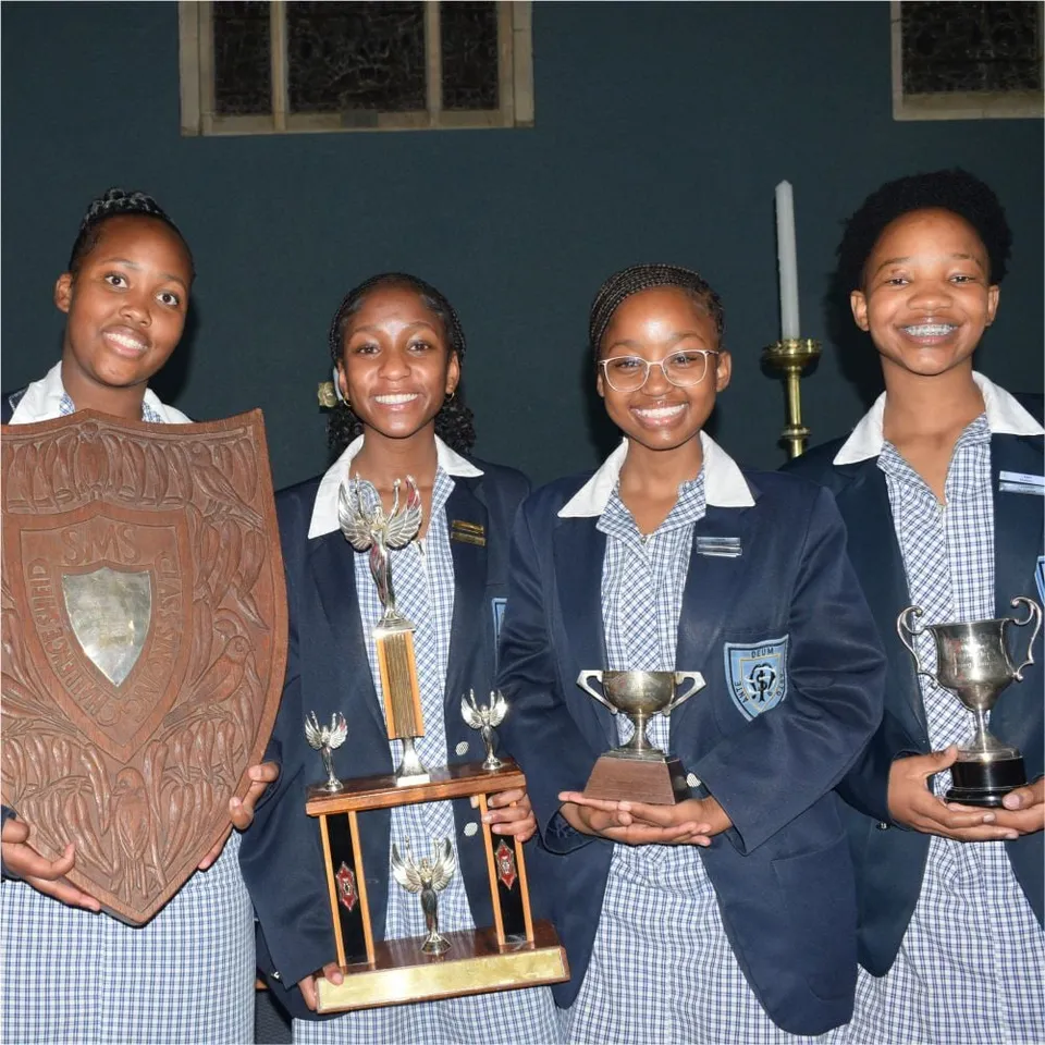 Quattro studentesse della St. Michael's School in uniforme sorridono mentre mostrano diversi trofei e uno scudo in legno.