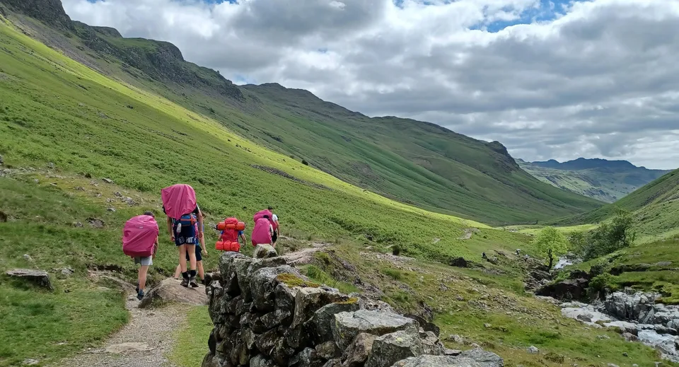 Un gruppo di studenti del Pangbourne College cammina su un sentiero roccioso in un paesaggio montano verde.