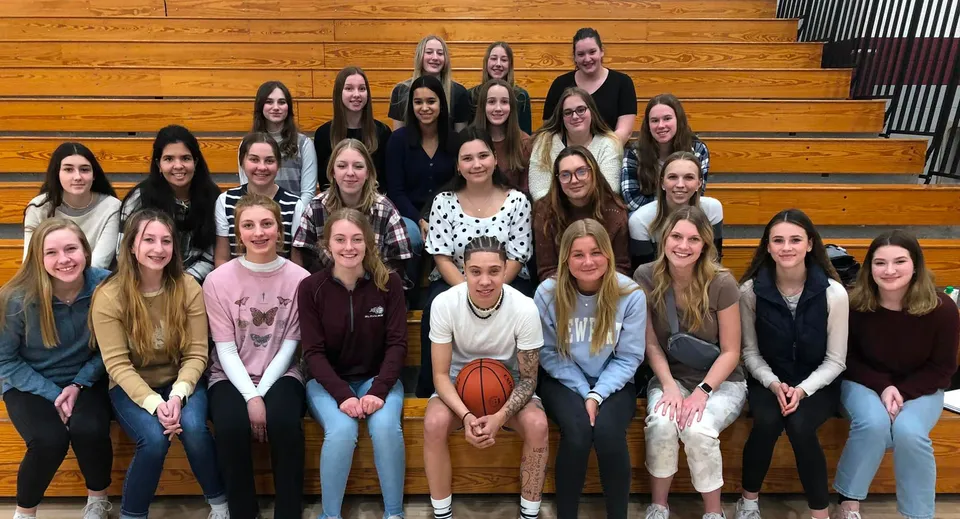 Studentesse della N.E.W. Lutheran High School sedute insieme su panchine di legno in palestra con un pallone da basket in primo piano.