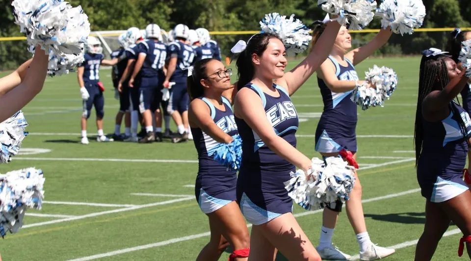 Le cheerleader della East Catholic High School si esibiscono sul campo da football con i giocatori sullo sfondo.
