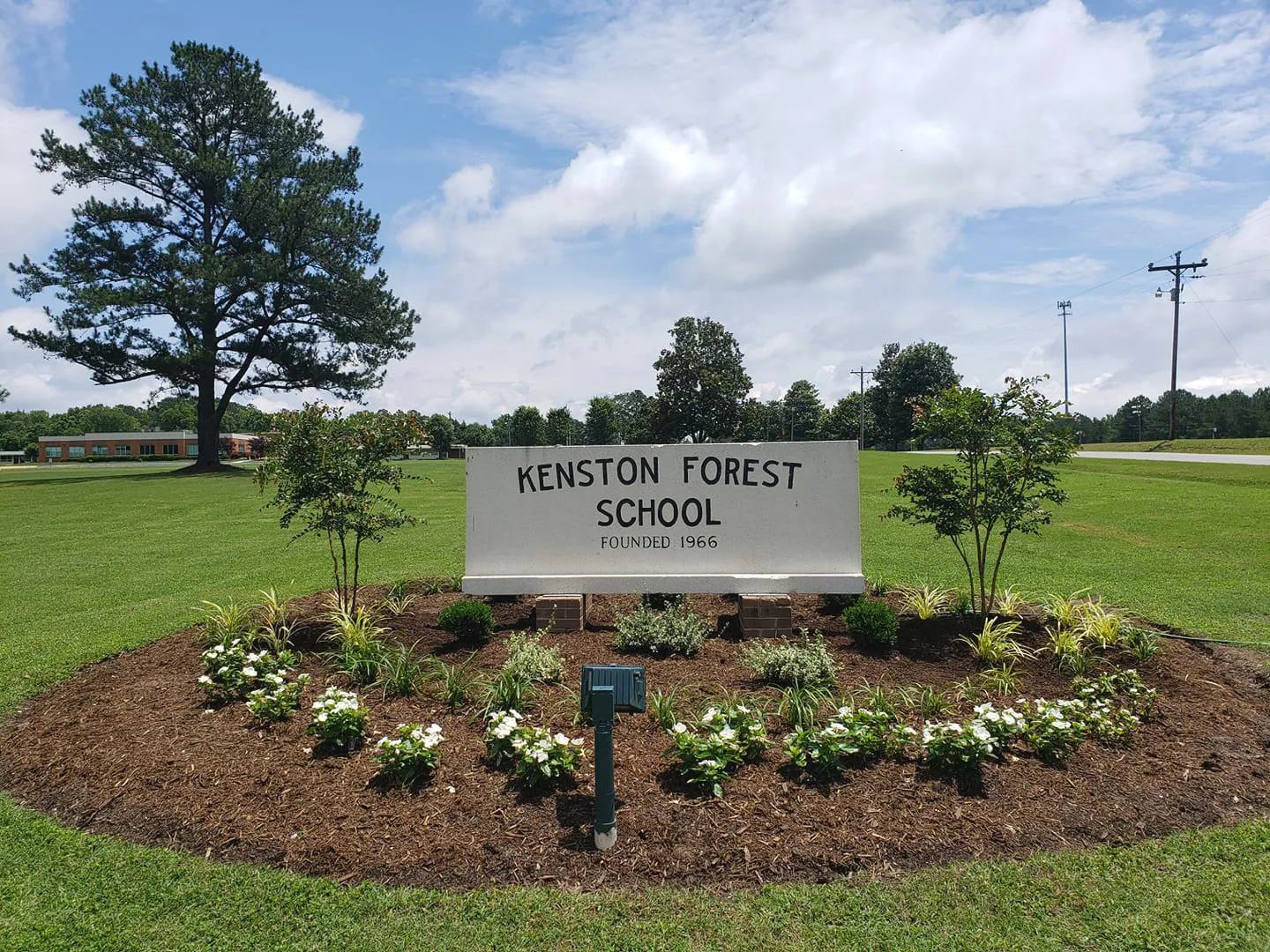 El cartel blanco de entrada de la Kenston Forest School en un césped bien cuidado con un parterre de flores.