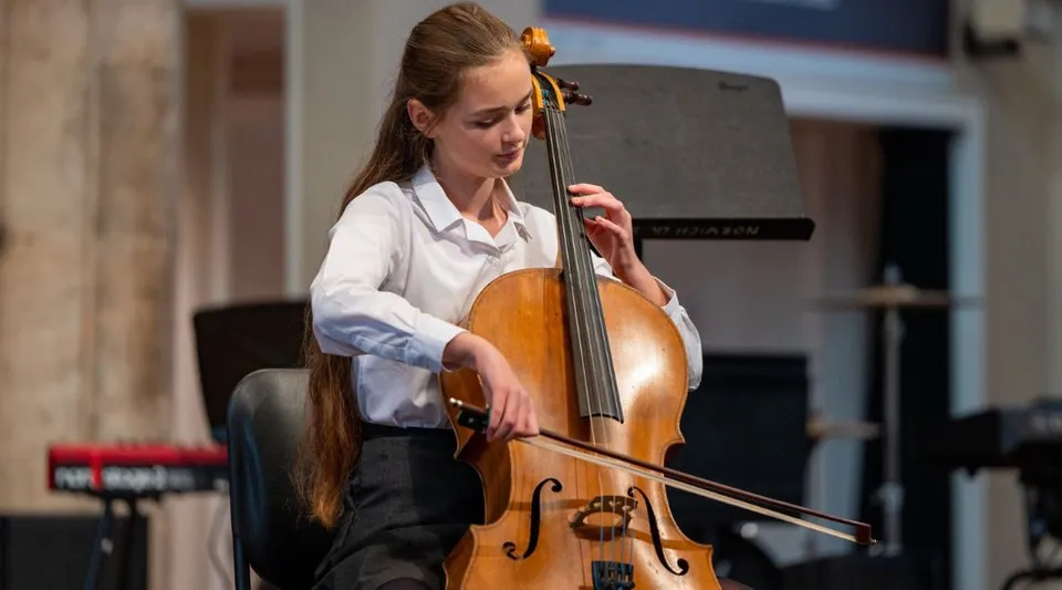 Una studentessa della Norwich School suona il violoncello in una stanza con pannelli di legno e strumenti musicali sullo sfondo.