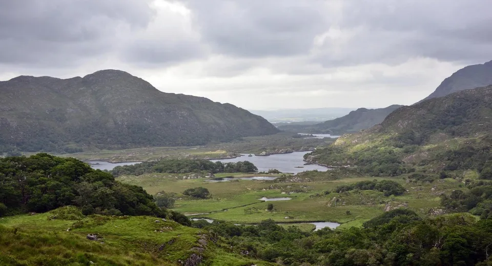 Una valle verde con un fiume sinuoso sotto un cielo nuvoloso nei pressi del Coláiste Choilm.