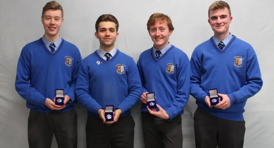 Quattro studenti del Summerhill College indossano divise blu e tengono in mano scatolette con anelli davanti a uno sfondo neutro.