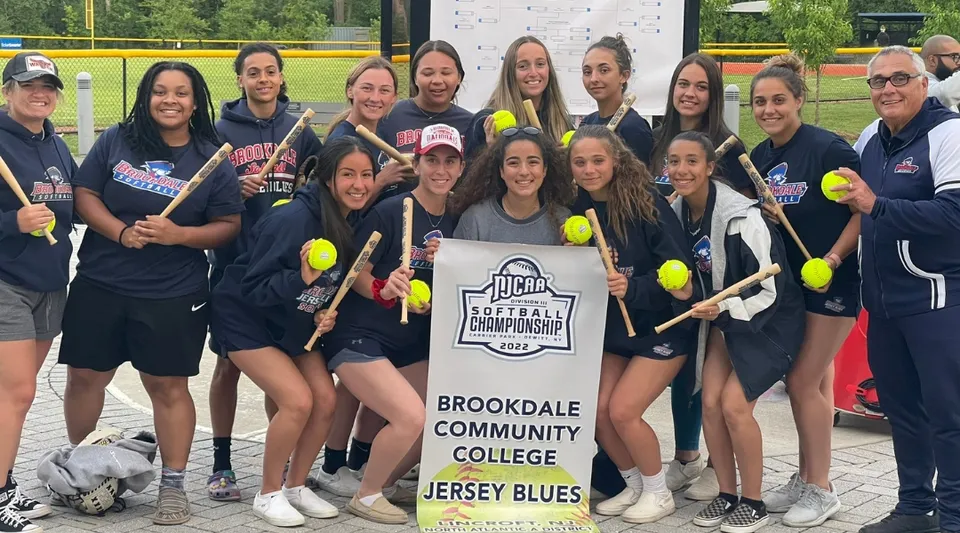 Un gruppo di giovani studentesse del Brookdale Community College posa con lo striscione della squadra di softball su un campo erboso.