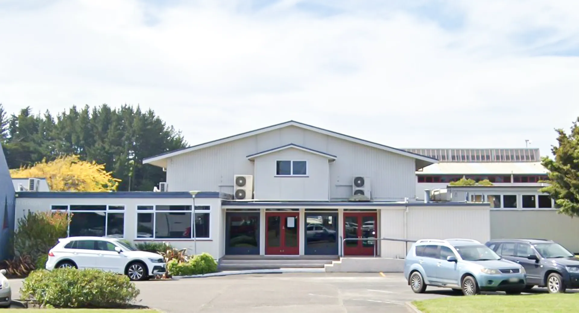 Il moderno edificio bianco con tetto obliquo nel campus del Manawatu College è circondato da veicoli parcheggiati e alberi.