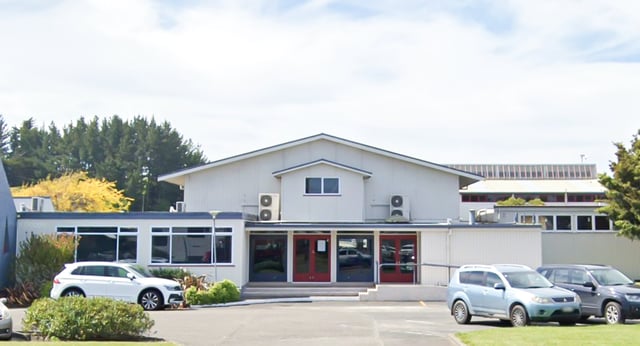 Il moderno edificio bianco con tetto obliquo nel campus del Manawatu College è circondato da veicoli parcheggiati e alberi.