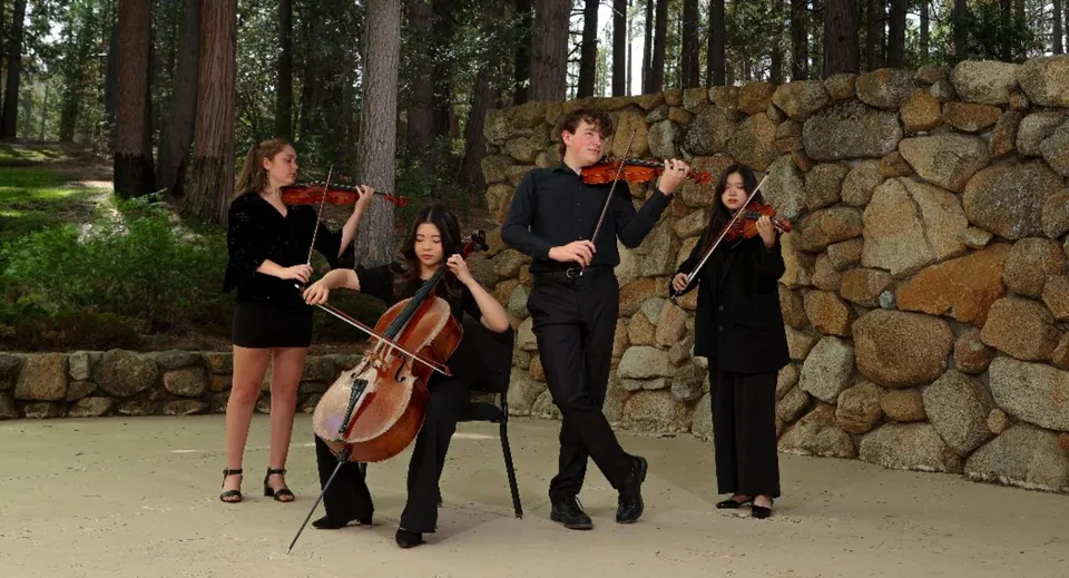 Quattro musicisti con strumenti ad arco si trovano nel campus della Idyllwild Arts Academy davanti a un muro di pietra nel bosco.