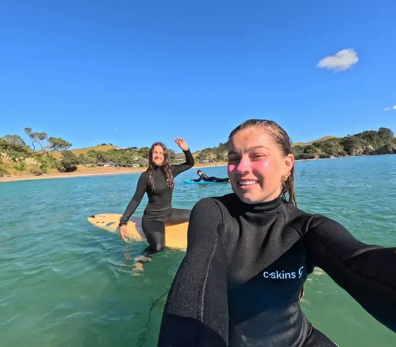Selfie de dos chicas con traje de neopreno y tabla de surf en el agua junto a la costa