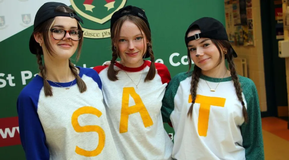 Tre studentesse del St. Patricks College in maglietta bianca SAT sono in piedi davanti a uno sfondo verde con stemma.