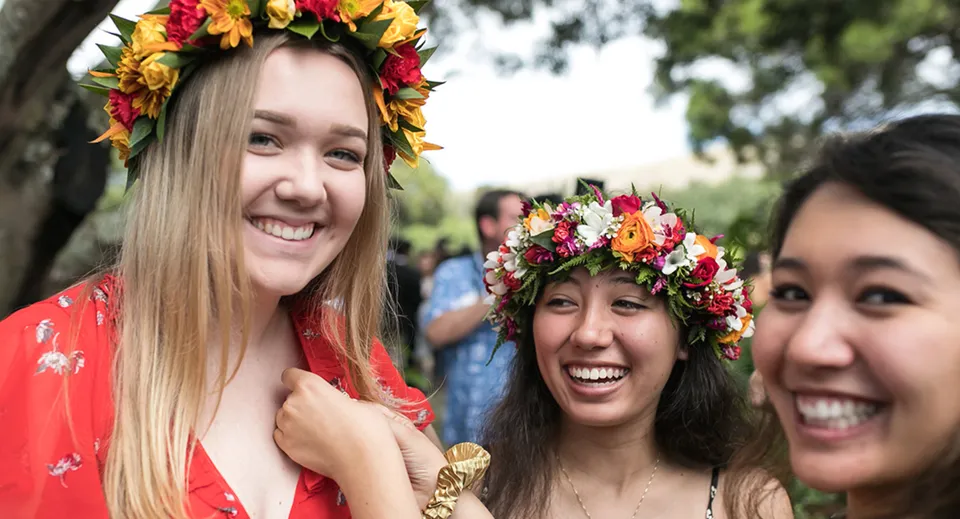 Tre studentesse della Hawaii Preparatory Academy indossano colorate corone di fiori e sorridono all'aperto davanti agli alberi.