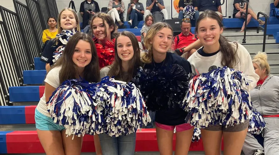 Un gruppo di cheerleader della Oak Mountain Academy posa in divise colorate con una folla sullo sfondo.