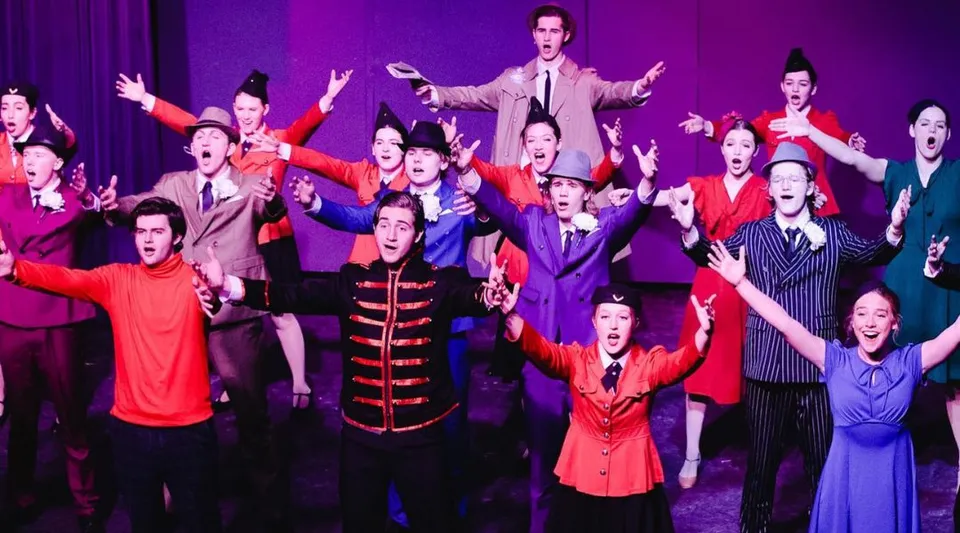 Gli studenti della Covenant Day School si esibiscono in un vivace spettacolo teatrale in costumi colorati con le braccia alzate.