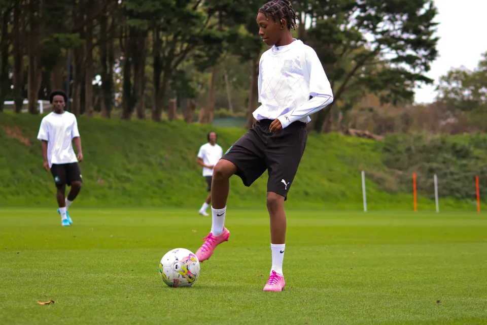 Uno studente del Plymouth College dribbla un pallone da calcio su un campo d'erba con altri giocatori sullo sfondo.