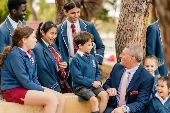 Estudiantes de la John Septimus Roe Anglican Community School con uniforme escolar conversan al aire libre con un adulto en traje.