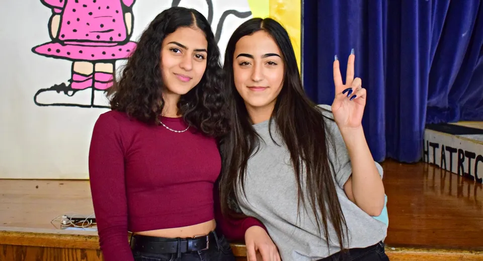 Due giovani donne con i capelli scuri posano davanti a un murale colorato nel campus dell'Honourable W.C. Kennedy Collegiate.