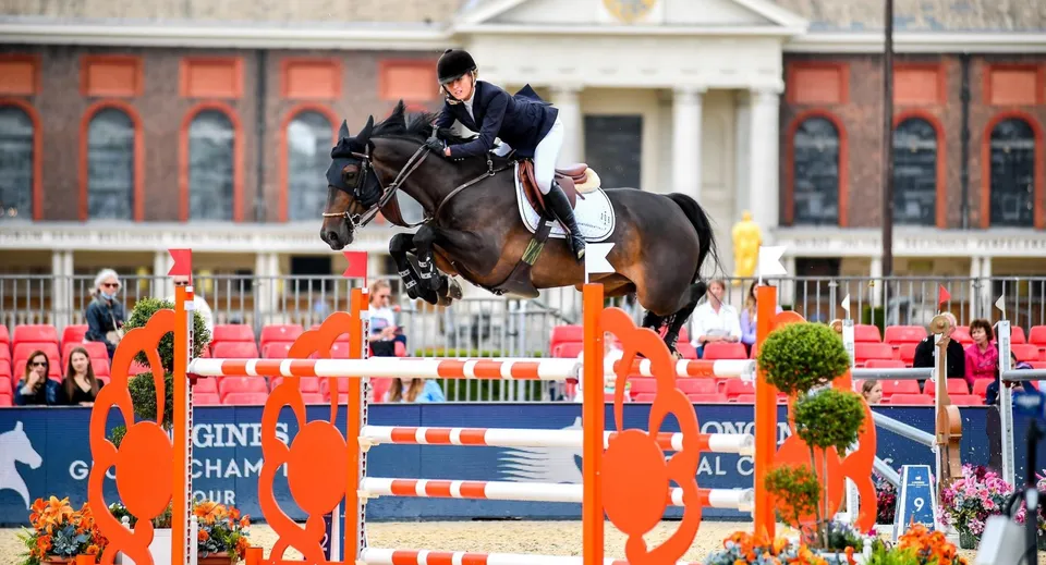 Un cavaliere salta un ostacolo arancione con un cavallo baio durante una competizione all'aperto nei terreni della Brentwood School.