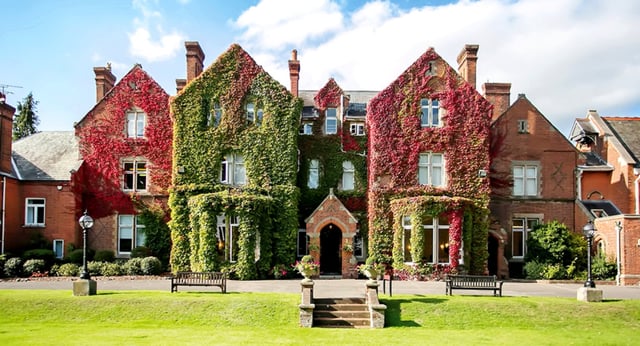 L'edificio storico in mattoni della Queen Anne’s School è circondato da fogliame rosso e verde e si trova dietro un prato curato con panchine.