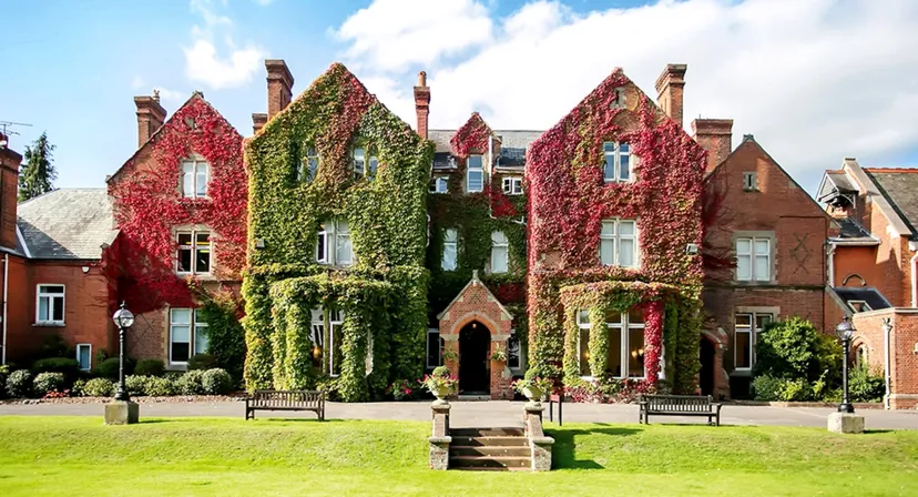 Un edificio storico in mattoni con un'architettura decorata è circondato dalle foglie rosse e verdi della Queen Anne’s School.