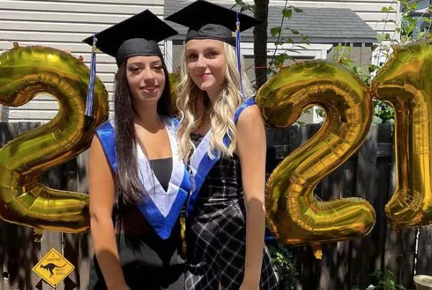 Dos alumnas de la A.Y. Jackson Secondary School posan con vestimenta de graduación ante globos dorados con forma de número con el "22".