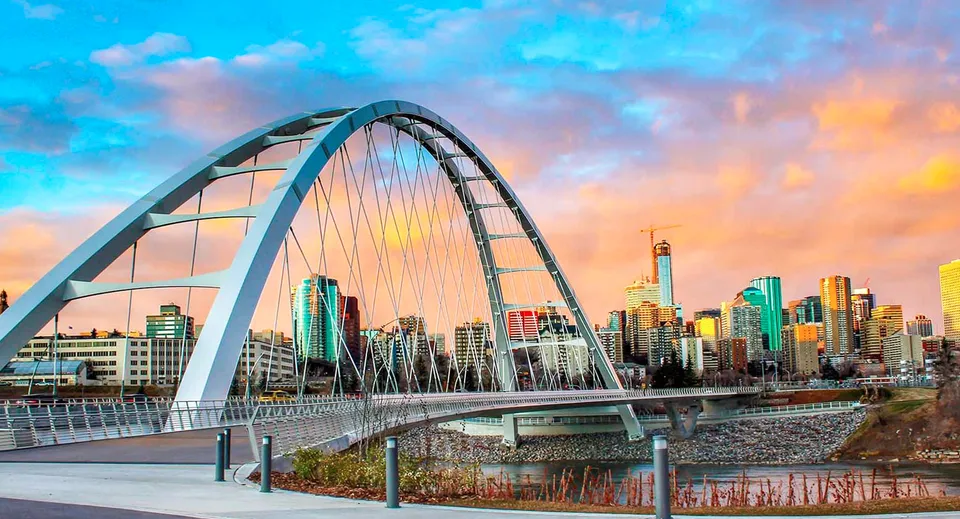 Un moderno ponte ad arco attraversa un fiume con un panorama della città dietro il campus della Queen Elizabeth High School al tramonto.