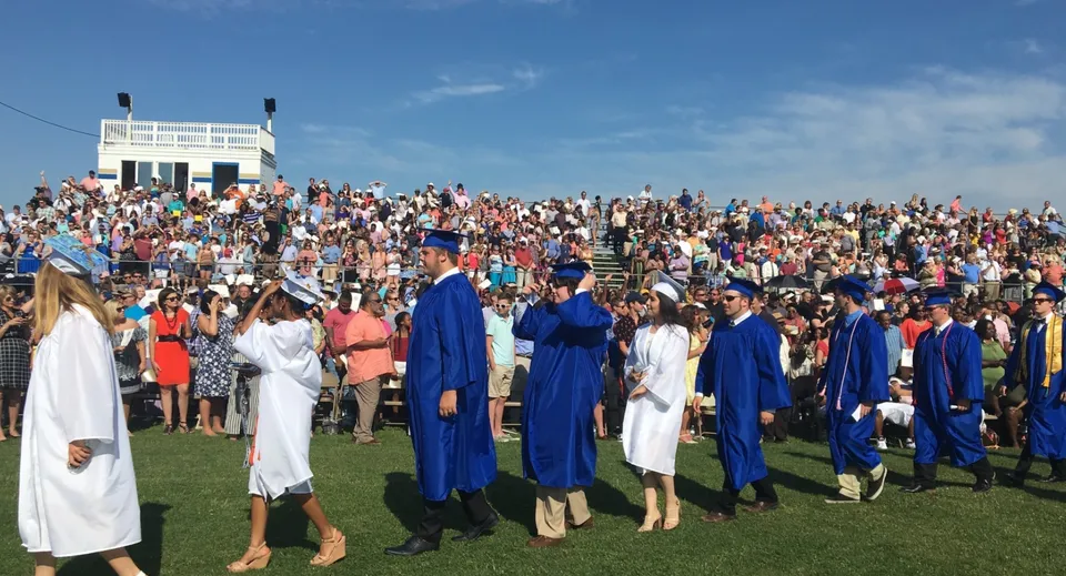 Un grande gruppo di diplomati della Smithfield State High School si trova su un prato con toga e tocco sotto un cielo blu.