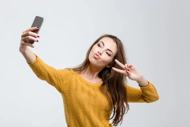 Junge Frau im gelben Pullover macht mit Kussmund und Peace-Zeichen ein Selfie
