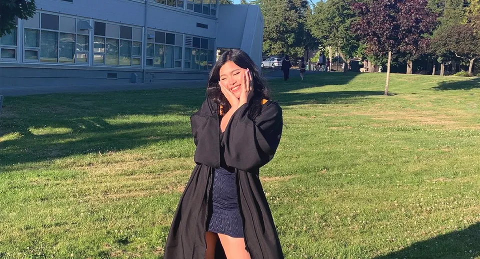 Una giovane donna in cappotto nero sta in piedi su un prato davanti all'edificio della Mount Douglas Secondary School.