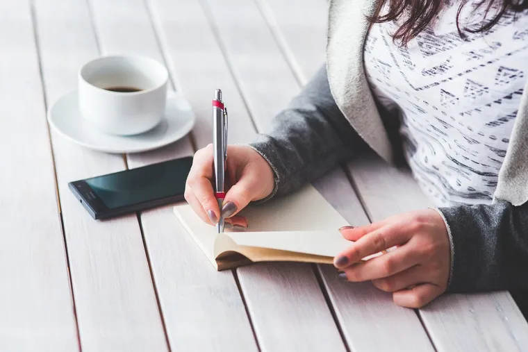 Hand schreibt mit Kugelschreiber in ein Notizbuch neben Kaffeetasse und Smartphone