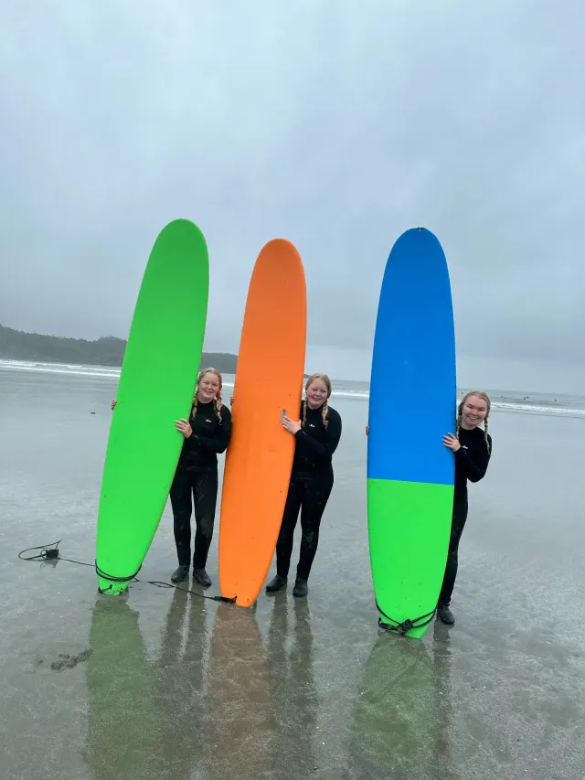 Tre studentesse in muta reggono lunghe tavole da surf colorate su una spiaggia canadese