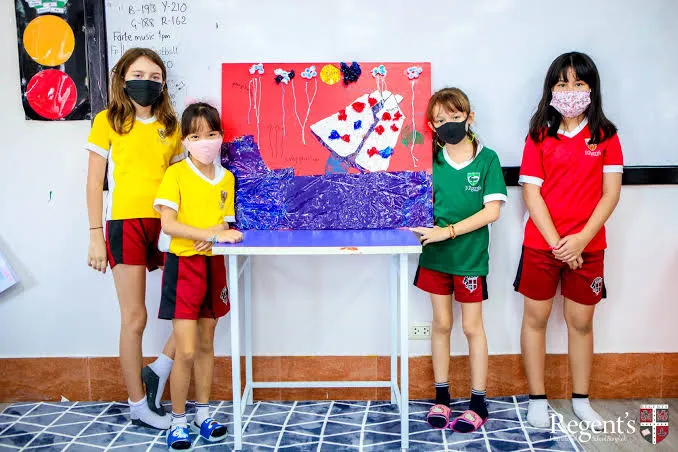 Vier Schülerinnen der Regent's International School Bangkok stehen mit Gesichtsmasken neben einem selbstgestalteten Plakat im Klassenzimmer.