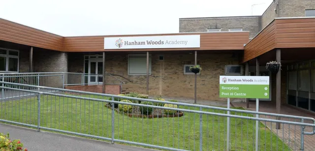 L'edificio in mattoni con facciata in legno della Hanham Woods Academy mostra il nome della scuola su un cartello accanto a una recinzione metallica e a un prato.