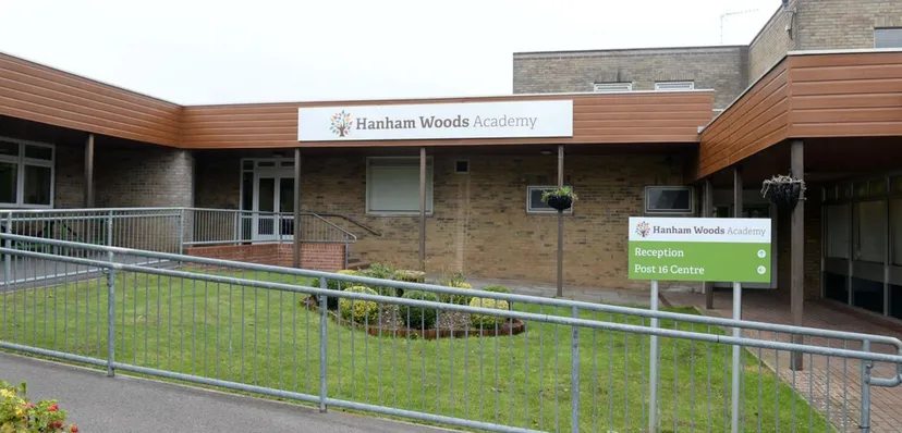L'edificio in mattoni con facciata in legno della Hanham Woods Academy mostra il nome della scuola su un cartello accanto a una recinzione metallica e a un prato.
