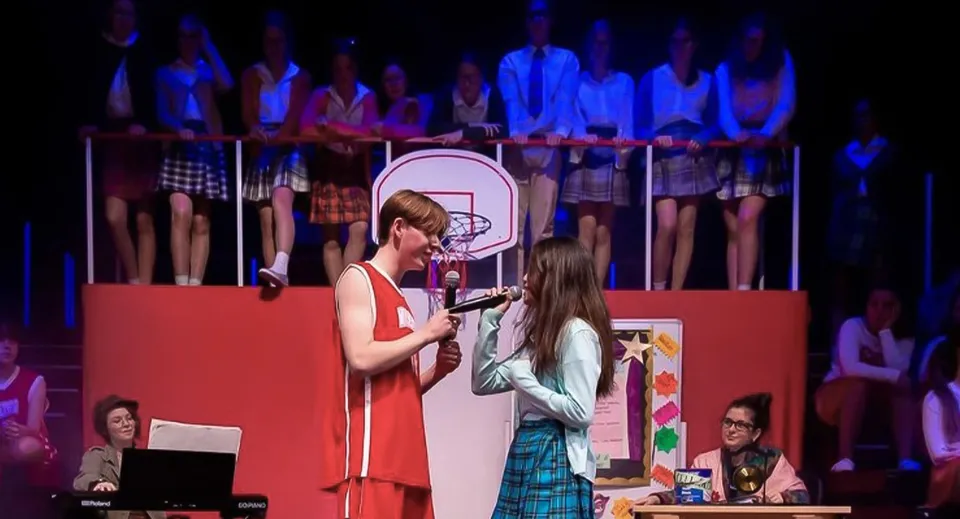 Una donna in abito rosso e una ragazza con la gonna a quadri sono sul palco del Malvern College durante una rappresentazione.