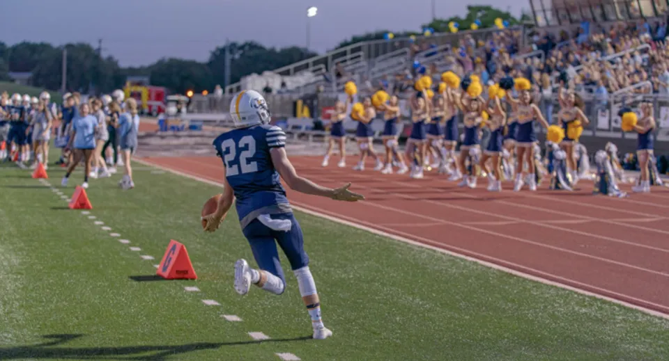 Un giocatore di football in maglia blu corre sul campo da gioco della St. John Paul II Catholic High School davanti agli spettatori sulle tribune.