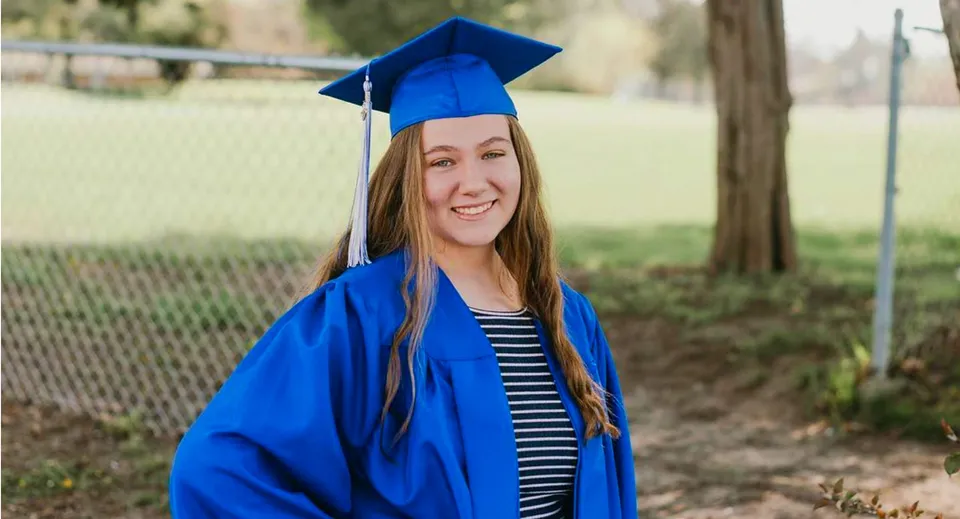 Una giovane diplomata in toga blu si trova nel campus della Christian Life School su un prato con alberi sullo sfondo.