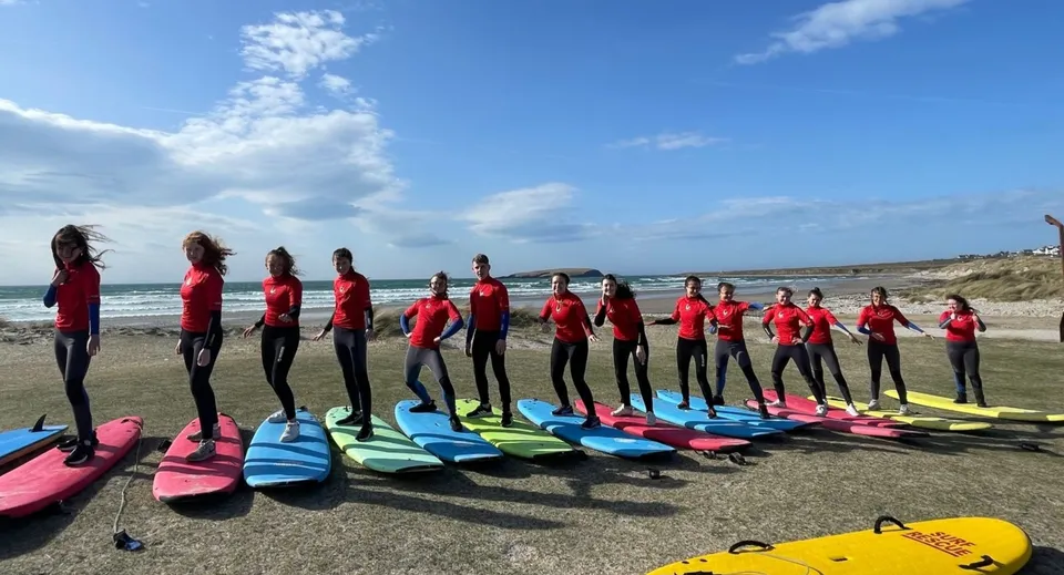Studenti del Coláiste Pobail Acla sono in piedi sulla spiaggia in muta rossa accanto a tavole da surf colorate con un paesaggio costiero sullo sfondo.