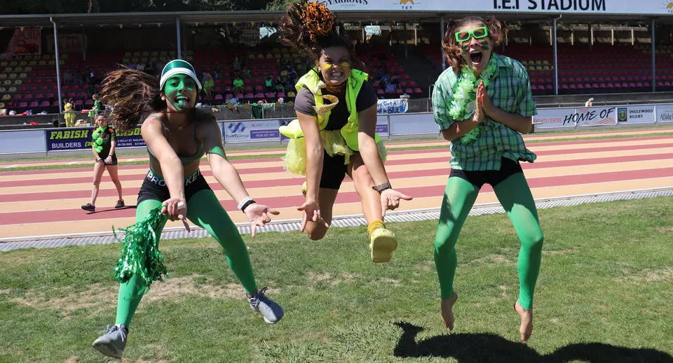 Studentesse della New Plymouth Girls’ High School posano con costumi colorati su un campo sportivo davanti allo stadio.