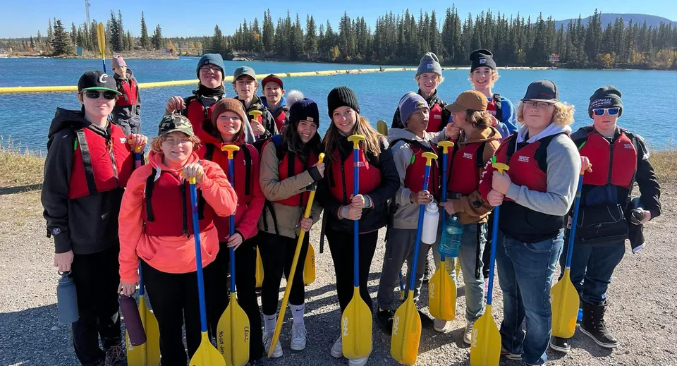 Un gruppo di studenti della Three Hills School si trova con giubbotti di salvataggio rossi e gialli davanti a un lago circondato da pini.