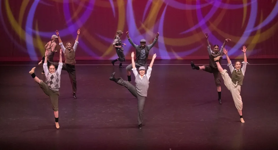 Un gruppo di ballerine e ballerini si esibisce sul palco del St. Andrew's College davanti a uno sfondo illuminato a colori.