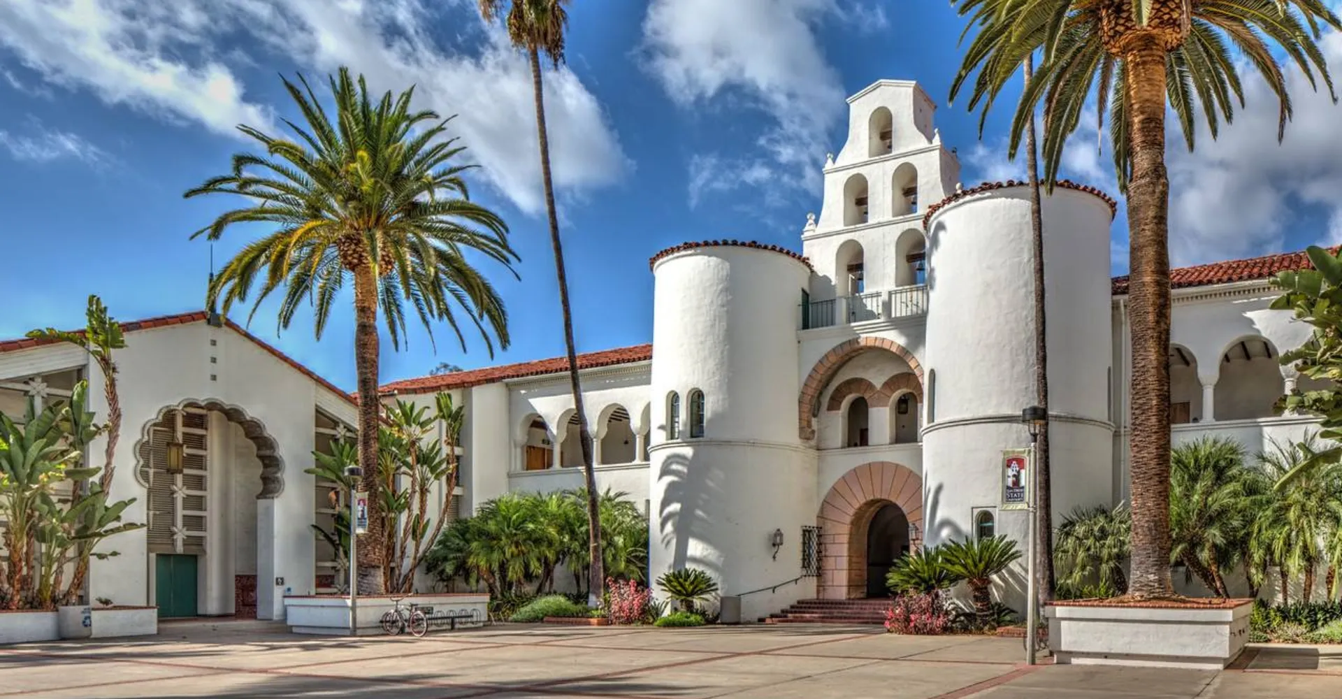 L'edificio bianco con torre campanaria della San Diego State University è circondato da palme e verde sotto un cielo azzurro.
